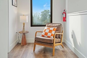 A corner room with a wooden chair, a small table with a lamp, a window with a view of trees, and a fire extinguisher on the wall.