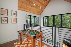 A modern interior with a foosball table, framed sports memorabilia, and large windows offering a view of greenery. - Vols-Themed Luxury Cabin | Indoor Pool | Sleeps 12 (Sevierville)