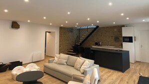 A modern living room with a sofa, a coffee table, and a kitchen area with a stone wall and a staircase. - Magnificent renovated farmhouse in peaceful countryside setting (Rives-du-Couesnon)