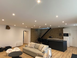 A modern living room with a sofa, a coffee table, and a kitchen area with a stone wall and a staircase. - Magnificent renovated farmhouse in peaceful countryside setting (Rives-du-Couesnon)