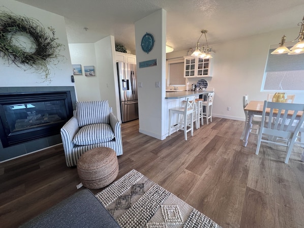 Een moderne woonkamer met een open haard, een gestreept fauteuil, een poef, en een eethoek met een houten tafel en stoelen.