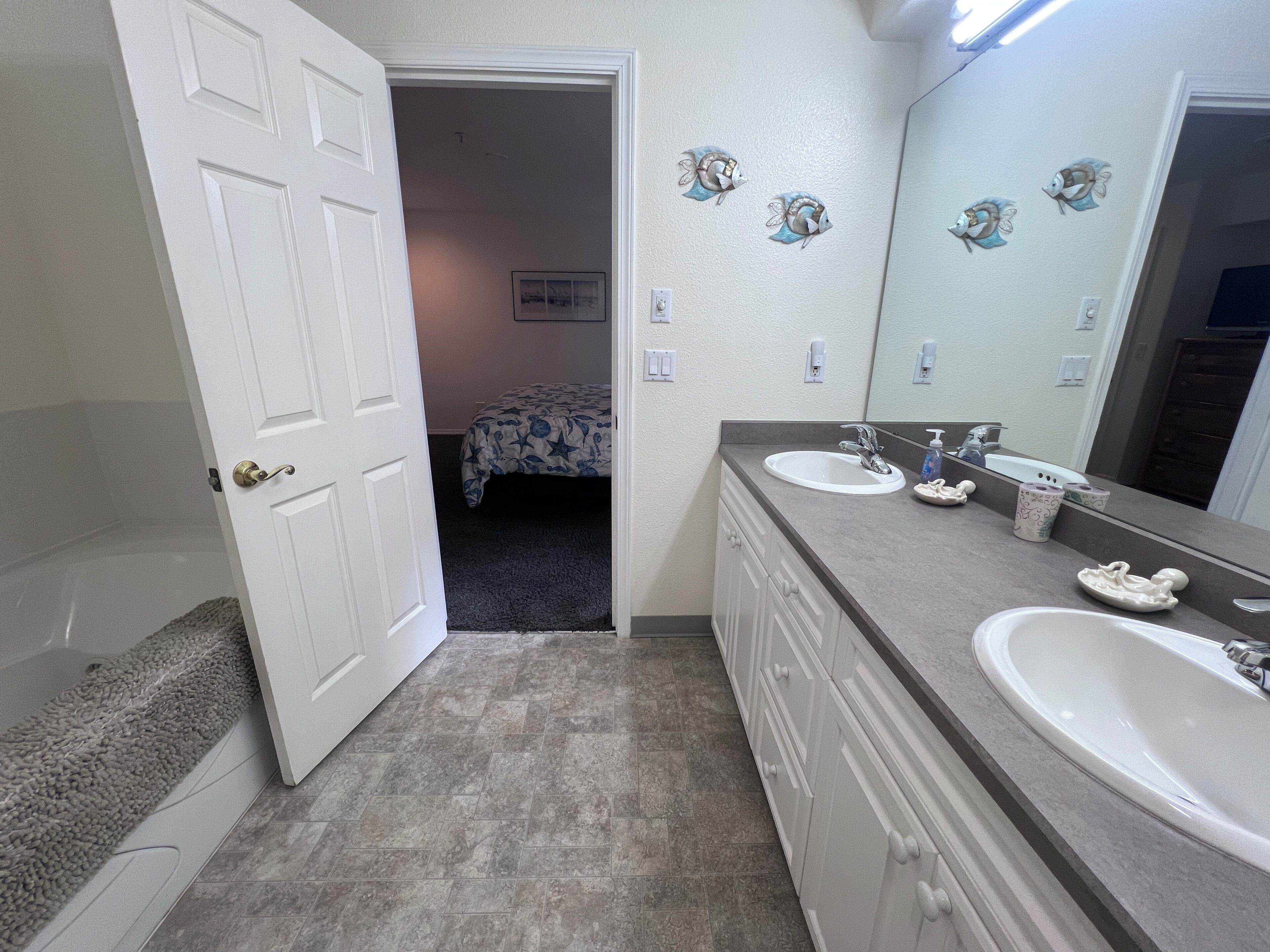 A bathroom with a bathtub, two sinks, a mirror, and a door leading to a bedroom.