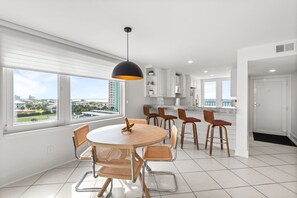 A modern kitchen with white cabinets, a dining area with a round table and chairs, and a large window offering a city view. - 🏖️ Santa Rosa Dunes 1061: Steps to the Gulf, Pool + Stunning Sound Views! (Pensacola Beach)