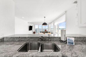 A modern kitchen with granite countertops, stainless steel sinks, and a pendant light. - 🏖️ Santa Rosa Dunes 1061: Steps to the Gulf, Pool + Stunning Sound Views! (Pensacola Beach)