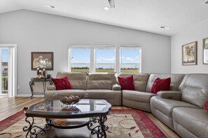 A spacious living room with a large L-shaped beige leather sofa, a glass coffee table, and a view of the outdoors through large windows.