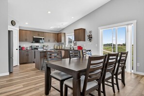 A modern kitchen with wooden cabinets, a dining area with a dark wooden table and chairs, and a large window with a view of the outdoors. - Sweet Esmeralda-FirePit, BBQ, Pets OK, Fenced Yard (Nampa)