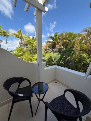 Ein Balkon mit zwei schwarzen Korbstühlen und einem kleinen Glastisch, umgeben von üppigen tropischen Pflanzen und wolkenlosem, blauem Himmel.