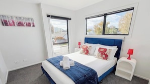 A bedroom with a bed, two windows, a nightstand, a lamp, and a wall art piece. - Relaxing Modern Stay in Upper Hutt Central (Upper Hutt)