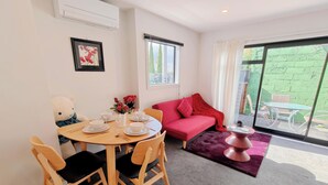 A dining area with a table set for two, a red sofa, and a large window with a view of greenery. - Relaxing Modern Stay in Upper Hutt Central (Upper Hutt)