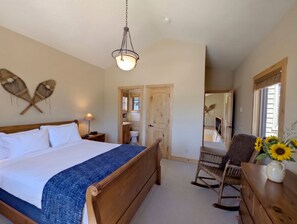 A bedroom with a wooden bed, a hanging light fixture, a rocking chair, and a view of the bathroom. - Riverside 5BR Home: Scenic Serenity & Trail Bliss (Keystone)