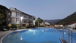 A swimming pool with lounge chairs and umbrellas, surrounded by buildings with balconies and a mountainous backdrop.