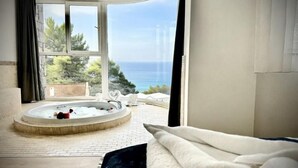 A bedroom with a large window, a circular hot tub, and a view of the sea.