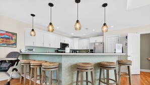 Une cuisine avec des meubles blancs, un îlot central doté de tabourets, des luminaires suspendus et une poussette.