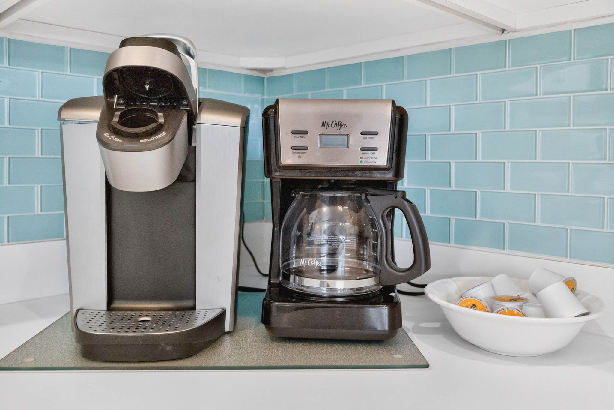 Eine Kaffeemaschine mit digitaler Anzeige, einem Glasbehälter und einer Schale mit Kaffeekapseln auf einer Arbeitsplatte.