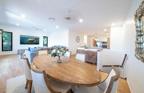 Eine moderne Küche mit einer zentralen Kochinsel, ein Essbereich mit Holztisch und ein Wohnzimmer mit Sofa und großem Fenster.