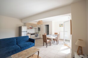 A modern living room with a blue sofa, a dining table with chairs, a kitchen in the background, and a lamp on a side table. - T3 ideal family with private garden, terrace, wifi and parking, 400 m from the lake (Arès)