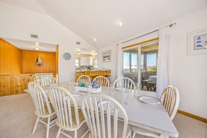 A dining area with a table set for six, white chairs, a view of the kitchen, and a large window with curtains. - Brand New Oceanside Salvo Home, Dog Friendly with Hot Tub (Salvo)
