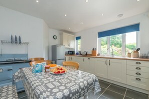 A kitchen with a dining table set for two, a refrigerator, and a window with a view of greenery. - Seaside Holiday Cottage Criccieth | Towyn (Criccieth)