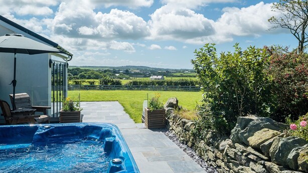 A swimming pool with a stone border, a patio area with seating, and a view of a green landscape. - Spacious Four Bedroom Holiday Accommodation On Anglesey | Bryndeusant (Holyhead, Ynys Môn)