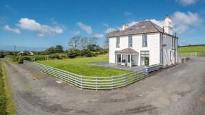 A white house with a covered patio, a gravel driveway, and a fenced lawn. - Spacious Four Bedroom Holiday Accommodation On Anglesey | Bryndeusant (Holyhead, Ynys Môn)