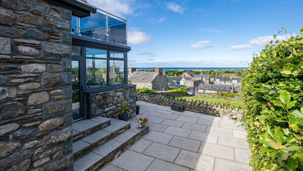 A stone building with a glass balcony, stone steps, and a view of the sea and town. - Modern Four Bedroom Property with Sea Views | Tan-Y-Foel (Dyffryn Ardudwy)