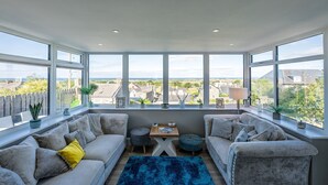 A spacious room with two grey sofas, a blue rug, and a small table with a lamp. Large windows offer a view of the surrounding area. - Modern Four Bedroom Property with Sea Views | Tan-Y-Foel (Dyffryn Ardudwy)