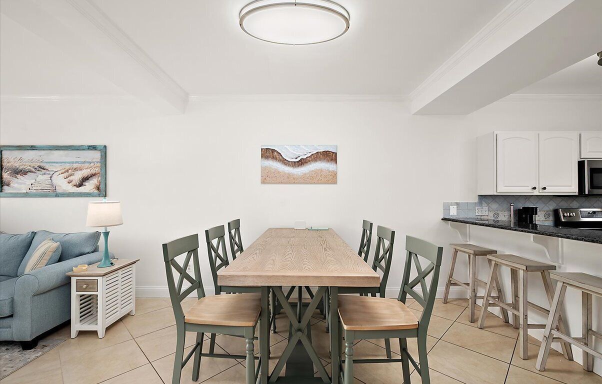 A dining area with a wooden table and chairs, a kitchen with white cabinets, and artwork on the walls.