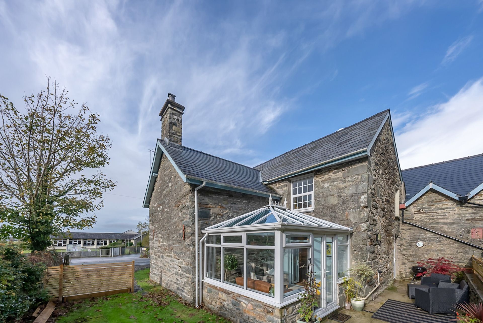 A stone house with a conservatory, a chimney, and a view of a school in the background.