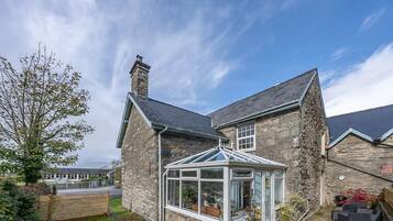 A stone house with a conservatory, a chimney, and a view of a school in the background.