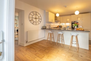 A modern kitchen with a central island, bar stools, a large wall clock, and white cabinetry. - Lovingly renovated three bedroom schoolhouse in Eryri | Isgoed (Talsarnau)
