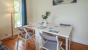 A dining area with a white table, two grey chairs, a vase with flowers, a painting of a beach scene, and a floor lamp. - Berkeley View (Looe)