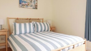 A bed with striped bedding, wooden headboard, bedside tables with lamps, and a painting on the wall. - Berkeley View (Looe)
