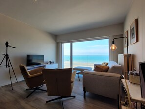 Ein modernes Wohnzimmer mit großem Fenster mit Blick auf den Strand, einem Fernseher und einem Stativ.
