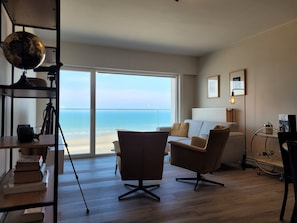 Ein modernes Wohnzimmer mit großem Fenster und Meerblick, inklusive Sofa, Schreibtisch mit Lampe und Globus auf einem Regal.