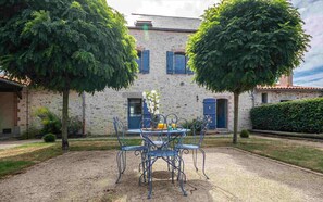 A courtyard with a table and chairs, surrounded by a stone building with blue shutters and a door. - 40 minutes from Le PUY DU FOU Gîte with swimming pool and private garden (Montaigu-Vendée)