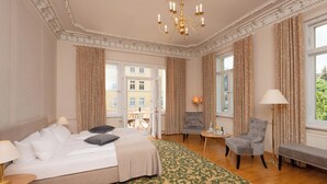 A hotel room with a bed, two armchairs, a bench, a small table, and a chandelier.
