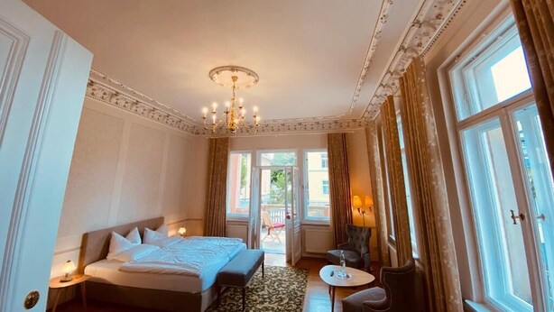A hotel room with a large bed, a small table, a chair, a chandelier, and a view of the outdoors.