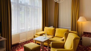 A hotel room with a yellow sofa, a small table with a bottle, a lamp, and a view through sheer curtains.