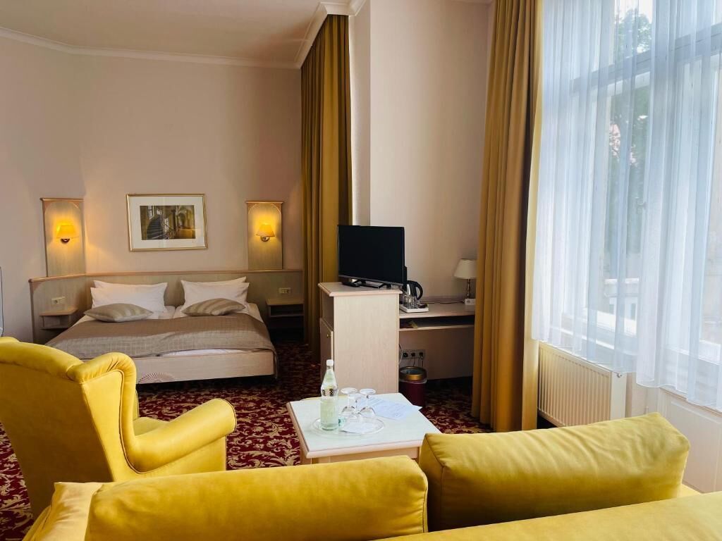 A hotel room with a bed, a desk with a computer, a chair, a table with a bottle, and a window with curtains.