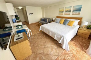 A hotel room with a bed, a desk with a chair, a small table with a plant, and a view of the beach through the window. - NEW! Fantastic Ocean Front Studio in Daytona Beach! (Daytona Beach)