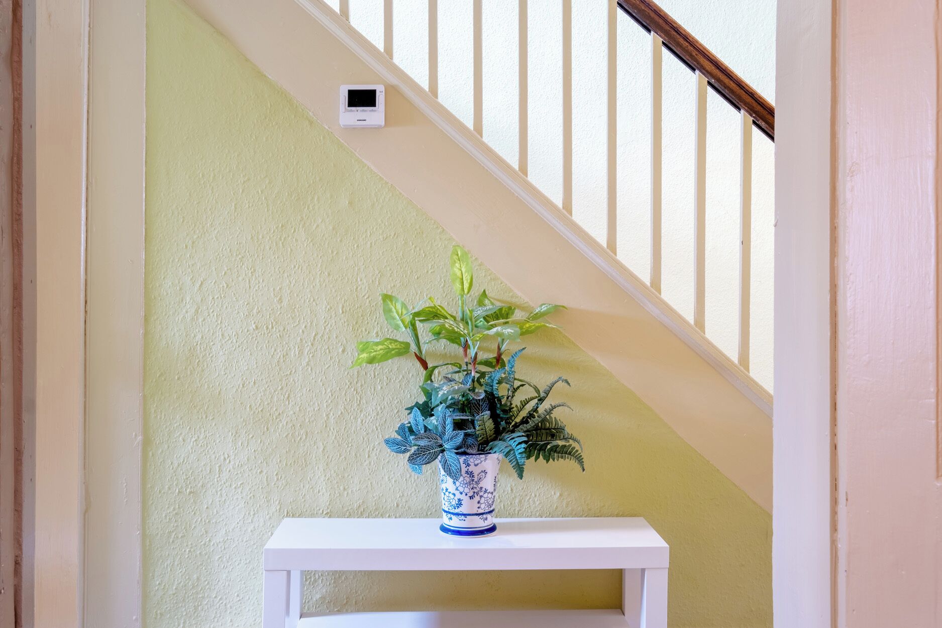 Un escalier avec une rampe blanche, un mur blanc et une étagère blanche sur laquelle se trouve un vase bleu et blanc orné de motifs et contenant une plante.