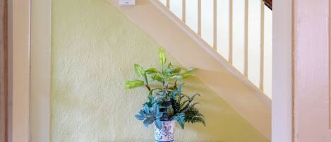 Una escalera con barandilla blanca, pared blanca y estante blanco con un jarrón azul y blanco con motivos geométricos que contiene una planta.