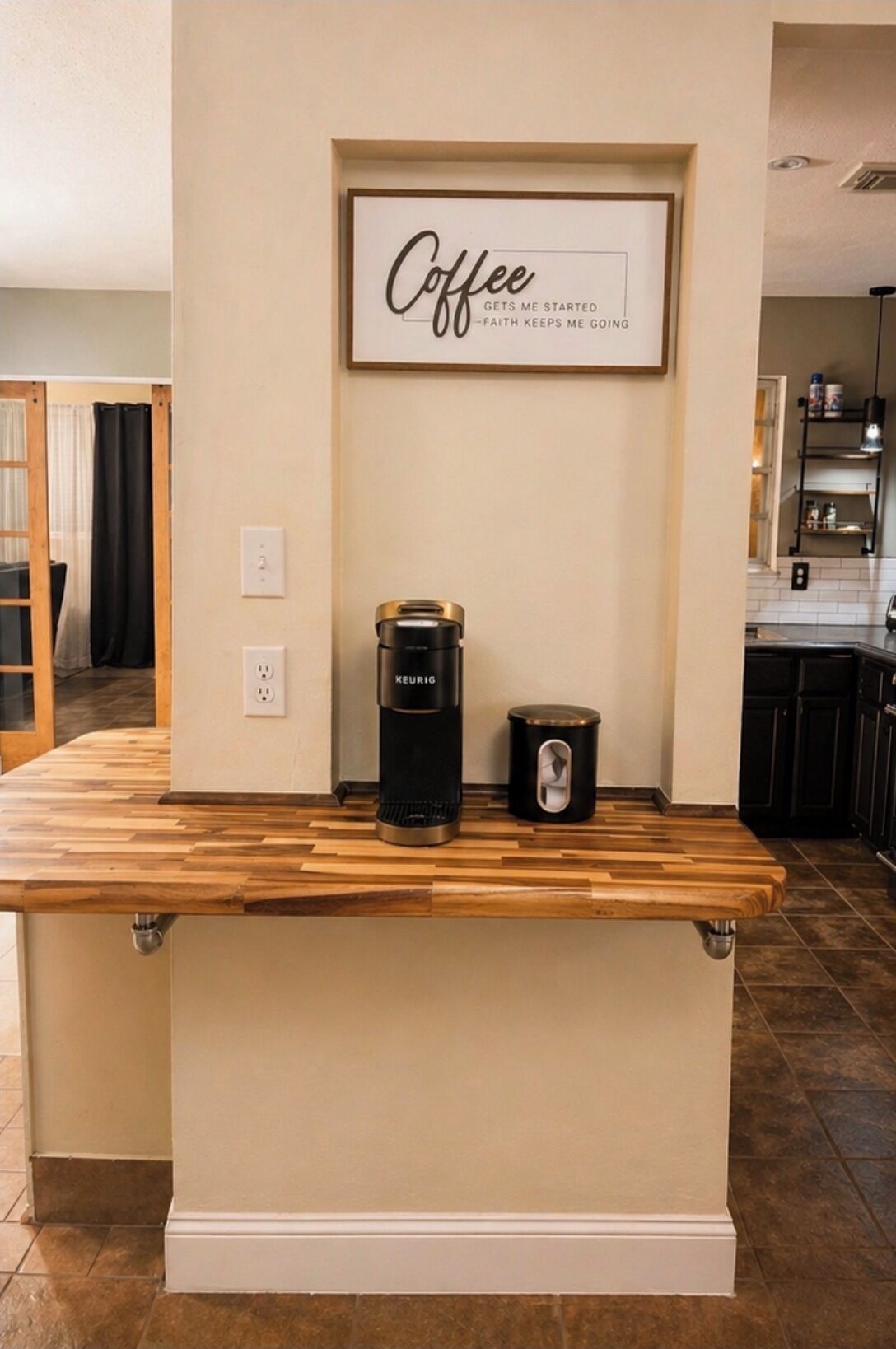Un coin cuisine avec une machine à café et une poubelle sur un plan de travail en bois, une pancarte encadrée au-dessus, et une vue sur une autre pièce.