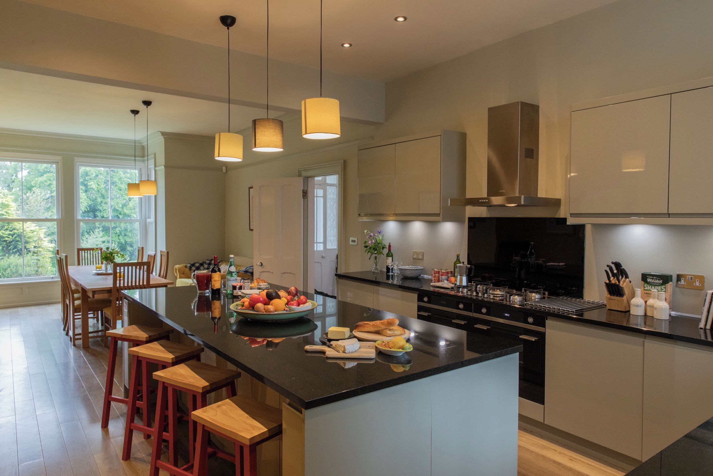 A modern kitchen with a large island, wooden bar stools, pendant lighting, and a view of the outdoors.