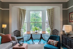 Ein Zimmer mit Erkerfenster, einem Esstisch, zwei Stühlen, einer Couch, einem kleinen Tisch mit Lampe und einem Schrank.