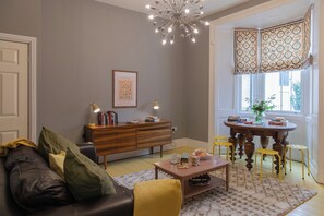 A living room with a sofa, coffee table, and a wooden cabinet. There is a window with curtains and a chandelier. - The Clarence (Bishop Auckland)