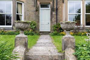 A stone pathway leading to a white door, flanked by two stone planters with greenery. - The Clarence (Bishop Auckland)