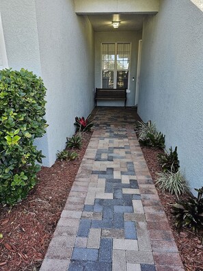 A paved pathway leading to a bench and a door, flanked by plants and mulch. - Magical landings Villa by Paradise Resort Vacations BRAND NEW TO THE RENTAL MARKET (Kissimmee)