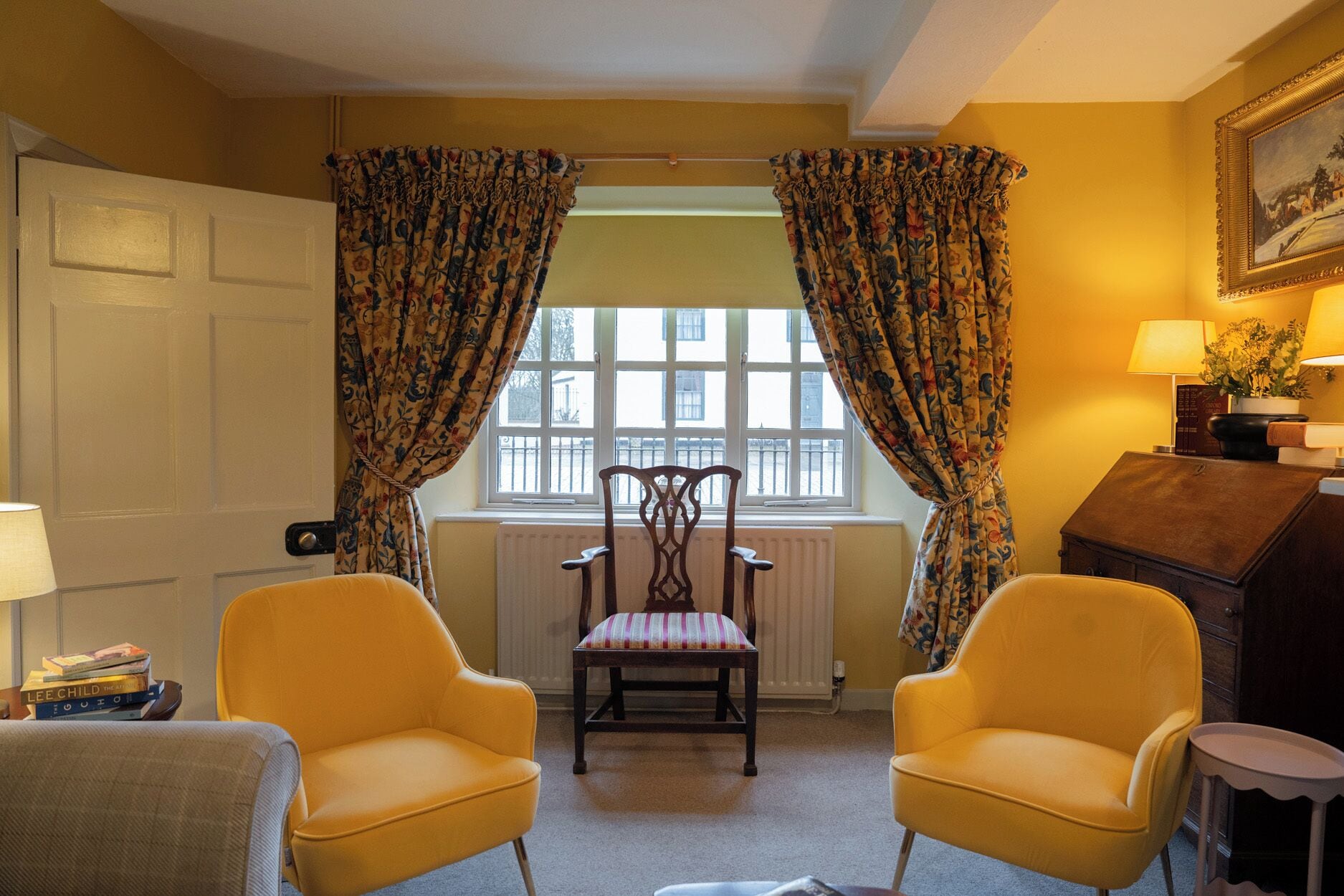 Een kamer met twee gele fauteuils, een houten stoel met een beklede zitting, een radiator, een raam met bloemen gordijnen, en een lamp op een tafel.