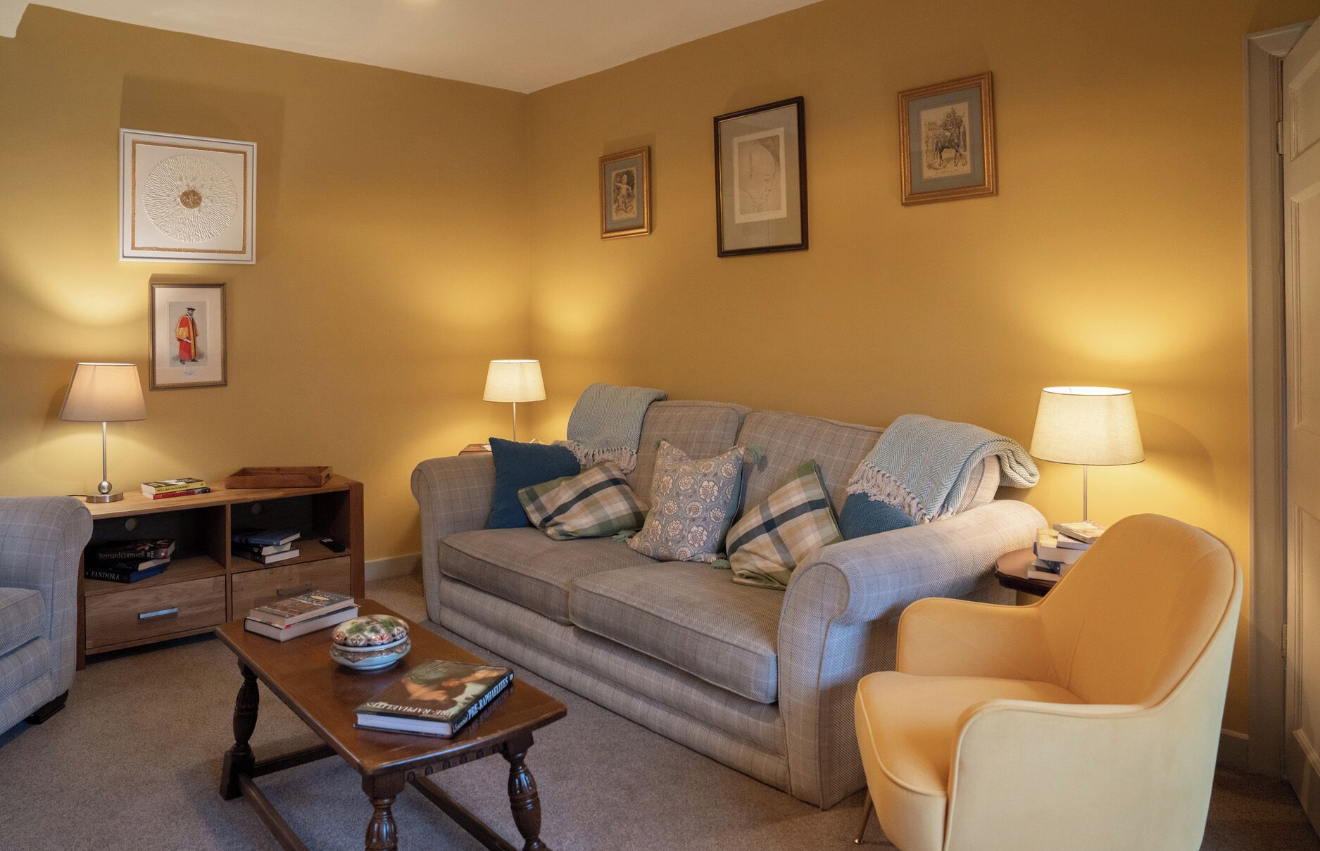 A living room with a sofa, armchairs, a coffee table, and lamps. There are framed pictures on the wall and a bookshelf with books.